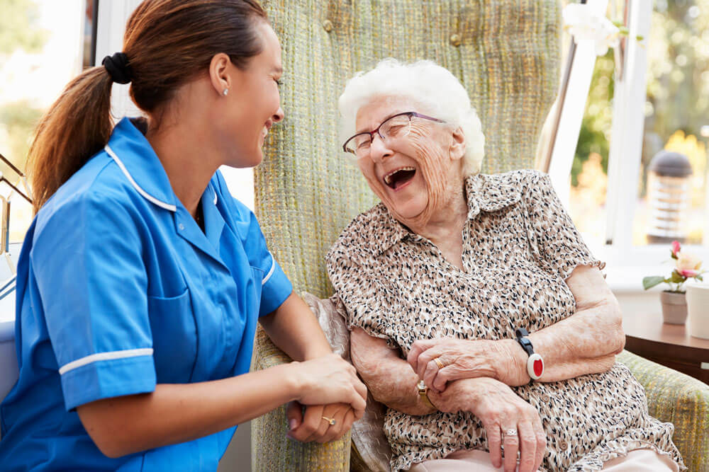 Ältere Frau sitzt im Stuhl und lacht mit Krankenschwester im Altersheim
