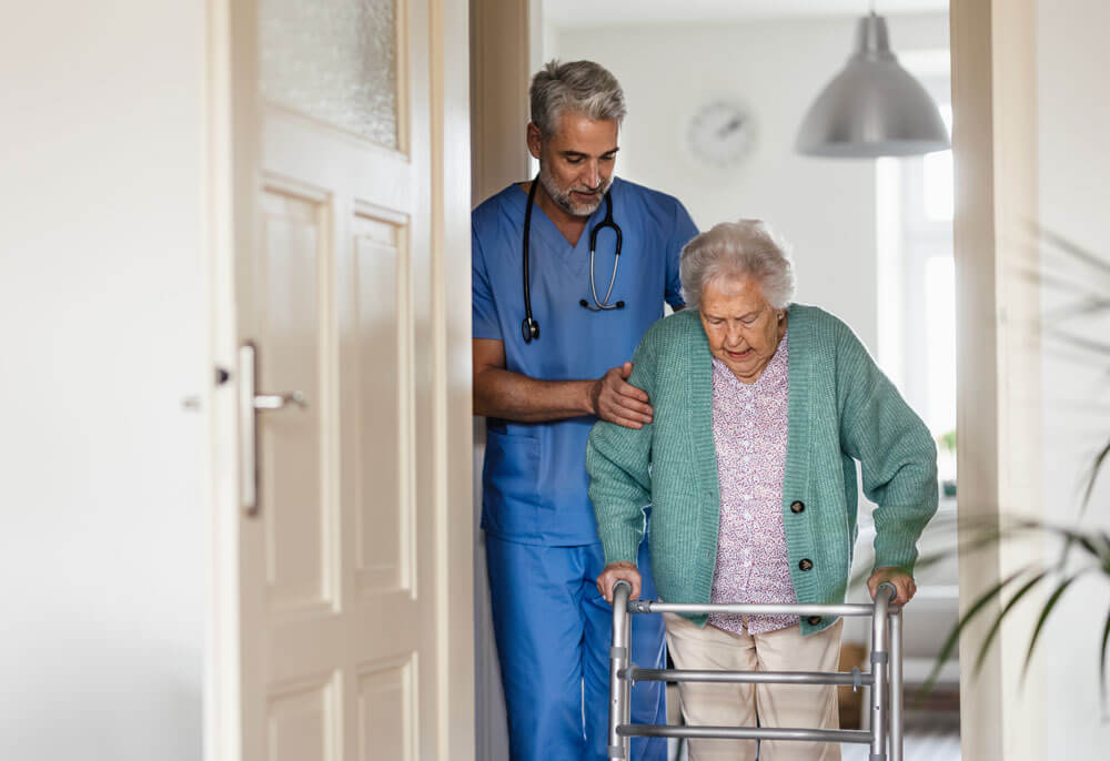 Betreuer hilft einer älteren Frau, mit einer Gehhilfe durch ihr Haus zu gehen.
